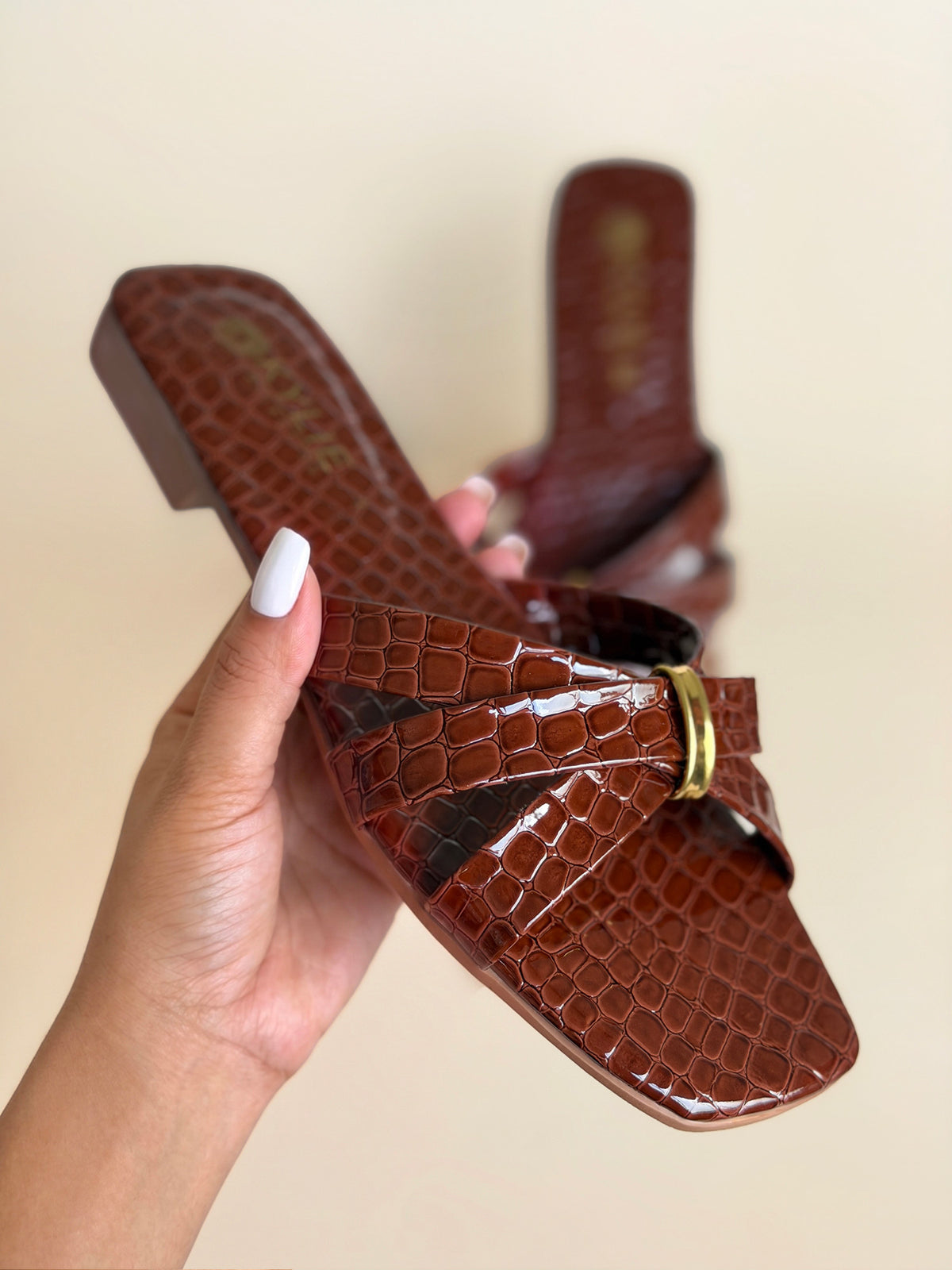 Close up of Kylie Collection ladies chestnut brown flat sandals shown on hand with crocodile texture crisscross straps and a small gold buckle detail in Sri Lanka