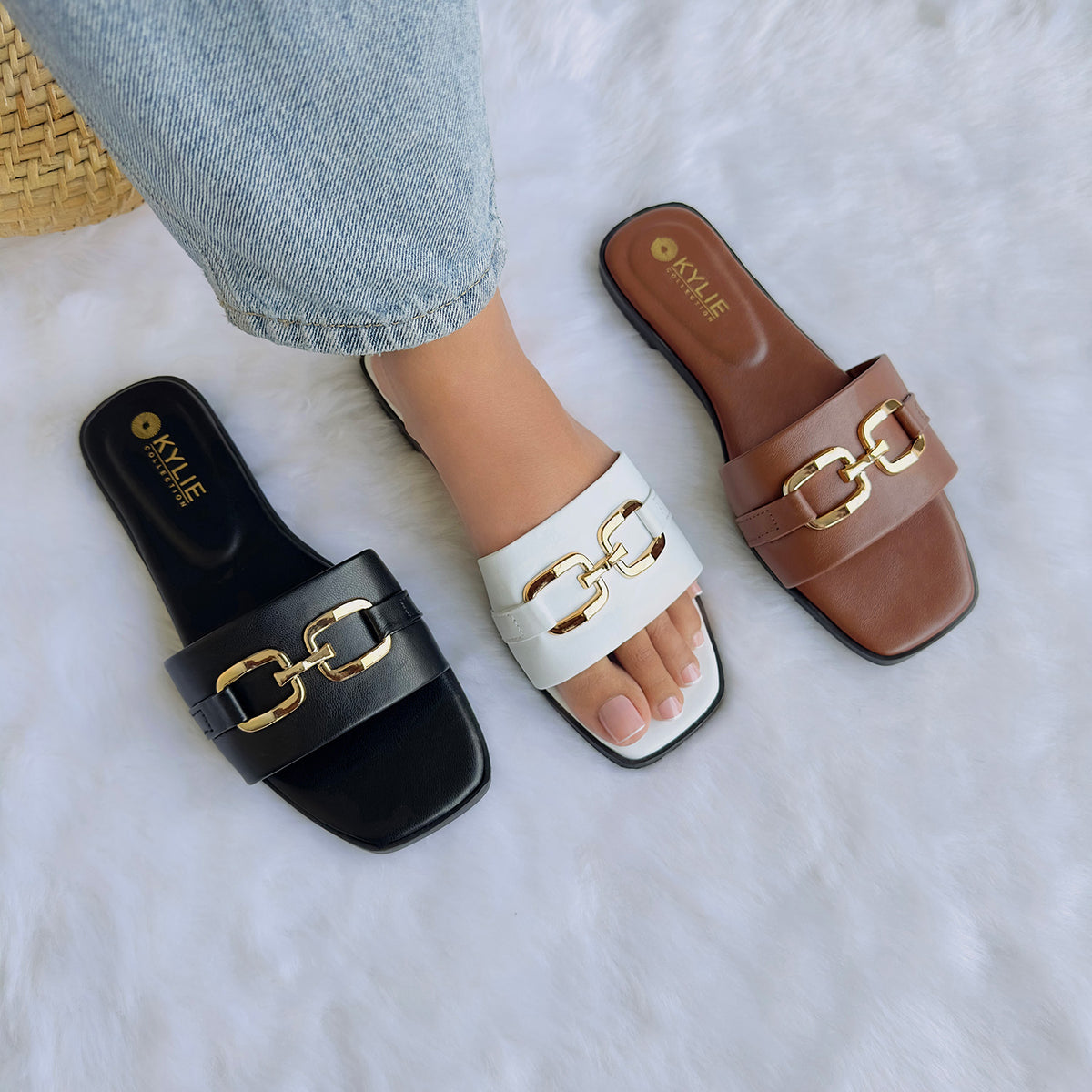 Three sandals (black, white, and brown) with gold chain detail on a fluffy white surface.