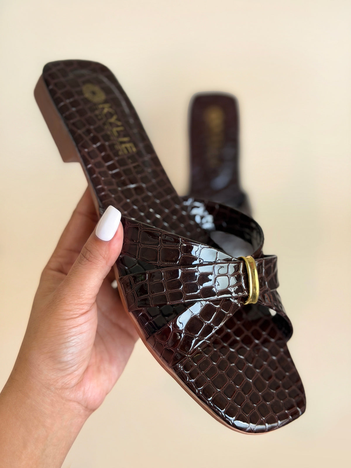 Close up of Kylie Collection ladies coffee brown flat sandals shown on hand with crocodile texture crisscross straps and a small gold buckle detail in Sri Lanka