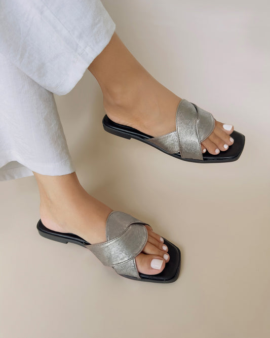 Close up of Kylie Collection ladies metallic silver slip-on flat sandals with wide cross straps and a black sole worn by a model in Sri Lanka