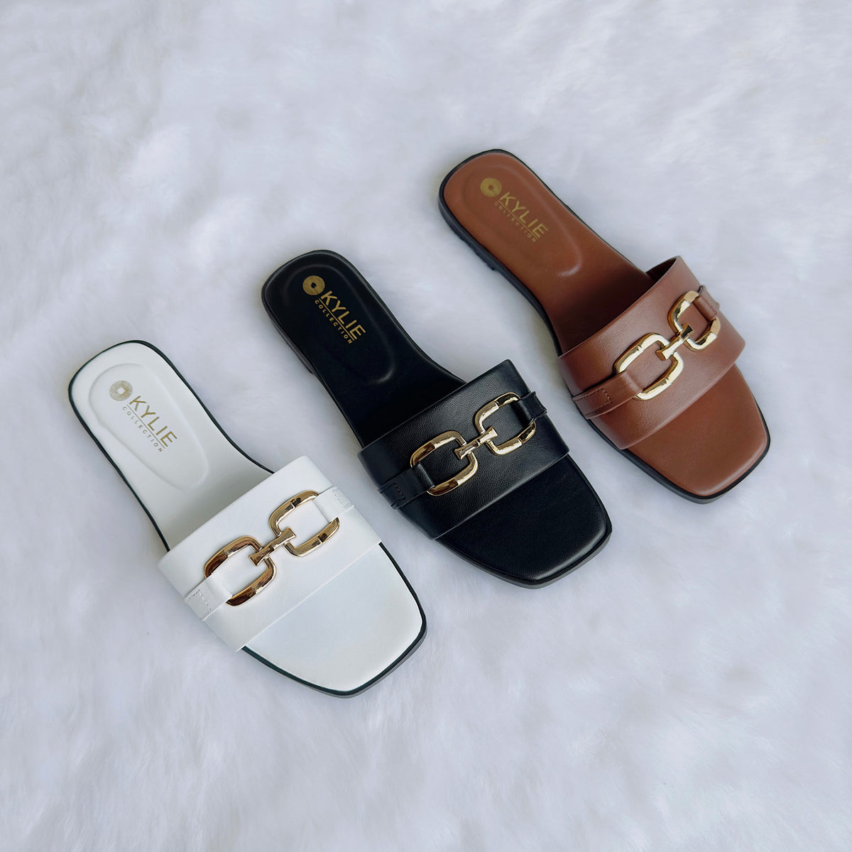 Three slide sandals in white, black, and brown colors, featuring a metal ornament on the strap